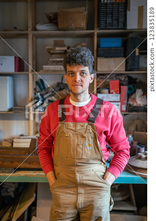Smiling man carpenter resting in workshop looking at camera putting hands in pockets leans on table Smiling man carpenter resting in workshop looking at camera putting hands in pockets leans on table 123835189