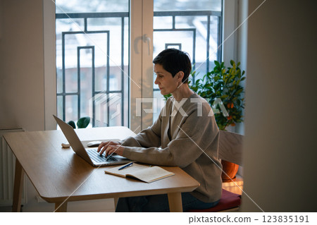 Tired focused woman works at laptop, take notes in notebook at home. Balances remote work and study Tired focused woman works at laptop, take notes in notebook at home. Balances remote work and study 123835191
