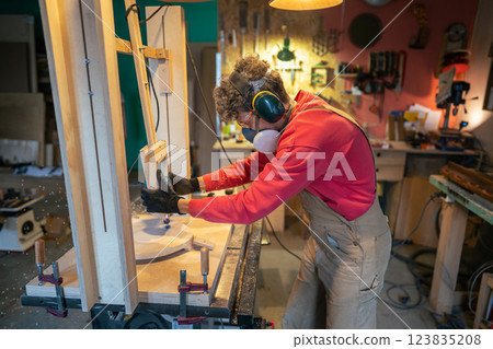 Carpenter in protective gear sands stool foundation in wood workshop. Smooth finishing, sculpting 123835208