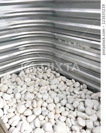 A close-up view of a corrugated metal window well with white decorative rocks at the base. The metal surface has minor scratches and wear, reflecting natural light. This functional design is commonly A close-up view of a corrugated metal window well with white decorative rocks at the base. The metal surface has minor scratches and wear, reflecting natural light. This functional design is commonly 123835239