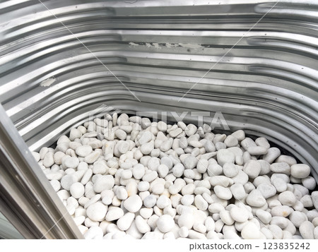 A close-up view of a corrugated metal window well with white decorative rocks at the base. The metal surface has minor scratches and wear, reflecting natural light. This functional design is commonly A close-up view of a corrugated metal window well with white decorative rocks at the base. The metal surface has minor scratches and wear, reflecting natural light. This functional design is commonly 123835242