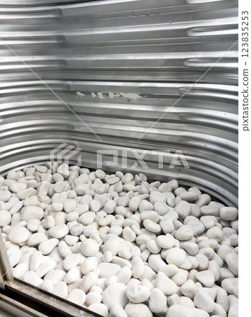 A close-up view of a corrugated metal window well with white decorative rocks at the base. The metal surface has minor scratches and wear, reflecting natural light. This functional design is commonly A close-up view of a corrugated metal window well with white decorative rocks at the base. The metal surface has minor scratches and wear, reflecting natural light. This functional design is commonly 123835253