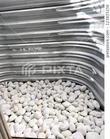 A close-up view of a corrugated metal window well with white decorative rocks at the base. The metal surface has minor scratches and wear, reflecting natural light. This functional design is commonly 123835256