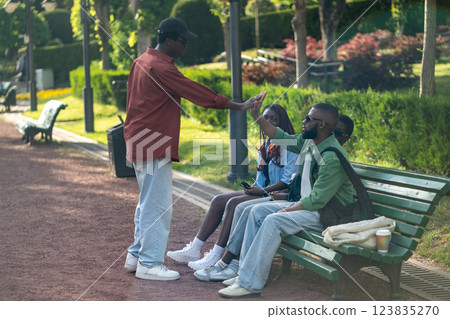 Park gathering, African American students greeting each other with high five, sit on bench together 123835270