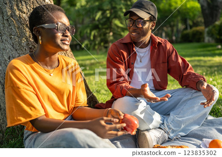 Black couple enjoys peaceful picnic, sit on blanket savoring snacks in conversation, date outdoors 123835302