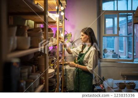 Woman places completed ceramic items on shelves in workshop of gift store, preparing for drying 123835336