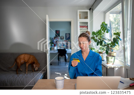 Pleased middle aged woman holding credit card sitting at table with laptop positive online shopping Pleased middle aged woman holding credit card sitting at table with laptop positive online shopping 123835475