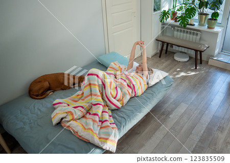 Happy woman lounging in bed with dog awakened from healthy sleep slowly waking up stretching arms. 123835509