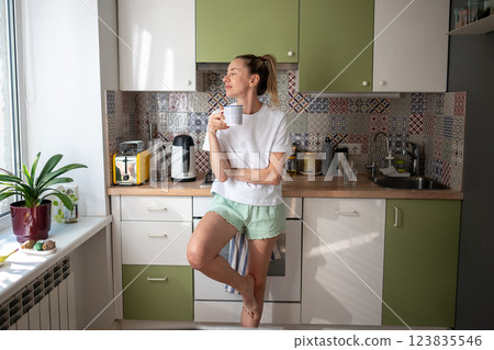 Satisfied happy woman standing alone in kitchen at cozy home atmosphere with cup of hot coffee. 123835546
