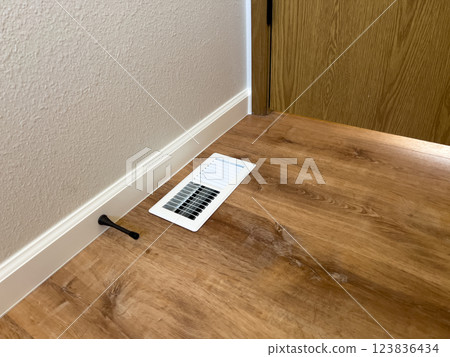Bathroom Floor Vent with Wood Flooring. High quality photo A close-up of a white air vent installed on a wood-look bathroom floor. The vent is positioned near the baseboard, complementing the newly 123836434