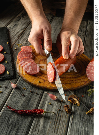 A person carefully slices cured sausage or salami on a wooden cutting board, surrounded by fresh spices and herbs. The warm light illuminates the rustic kitchen atmosphere 123836574