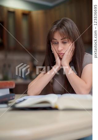 Adult woman with ADHD or dyslexia experiencing frustration and stress, sitting at desk with open book, holding her head in hands amidst study challenges 123836695