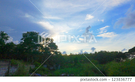 A serene landscape in West Surabaya, Indonesia, featuring lush greenery, scattered trees, a distant building, and a clear blue sky with a visible moon. 123836777