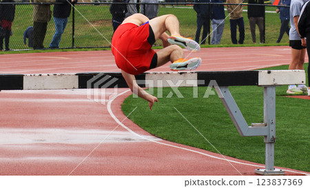 Runner Attempts a Hurdle Jump at a Track Meet During a Rainy Day While Spectators Observe From the Sidelines 123837369