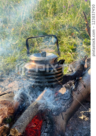 Close-up of metal kettle heating over campfire. Flames and smoke curl around well-used kettle, which rests on logs. Blur of green grass, emphasizing rustic outdoor setting on background. Close-up of metal kettle heating over campfire. Flames and smoke curl around well-used kettle, which rests on logs. Blur of green grass, emphasizing rustic outdoor setting on background. 123837450