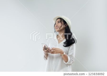 Beautiful young Asian woman tourist in straw hat standing posing with hand holding her smart phone and while looking at the copy space above her, woman advertising online purchase Beautiful young Asian woman tourist in straw hat standing posing with hand holding her smart phone and while looking at the copy space above her, woman advertising online purchase 123838092