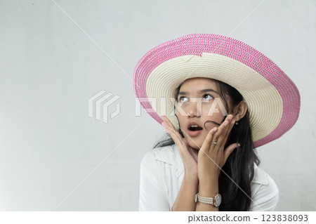 Young asian woman in white suit, pink striped straw hat with surprised expression towards the side on white background, showing surprise. Young asian woman in white suit, pink striped straw hat with surprised expression towards the side on white background, showing surprise. 123838093