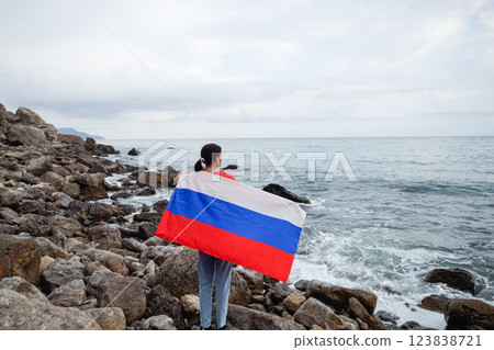 A brunette woman with a Russian flag on her shoulders stands on a rocky seashore. Independence day, patriotic holiday concept 123838721