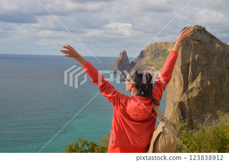 A tourist with a backpack travels through the Crimean village of Novy Svet, Cape Fiolent. Girl on the edge of a cliff enjoying the sea view. Active summer holiday, adventure lifestyle. A tourist with a backpack travels through the Crimean village of Novy Svet, Cape Fiolent. Girl on the edge of a cliff enjoying the sea view. Active summer holiday, adventure lifestyle. 123838912