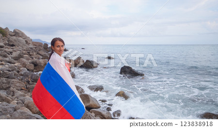 A brunette woman with a Russian flag on her shoulders stands on a rocky seashore. Independence day, patriotic holiday concept 123838918