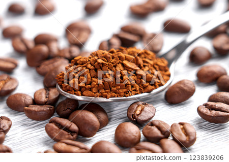 Metal spoon with instant coffee granules surrounded by whole roasted Arabica beans on white wooden surface. Macro shot.  123839206