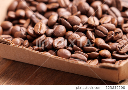 Light roast Arabica coffee beans in kraft paper box on wooden surface. Aromatic drink grains in cardboard packaging. Macro shot. Light roast Arabica coffee beans in kraft paper box on wooden surface. Aromatic drink grains in cardboard packaging. Macro shot. 123839208