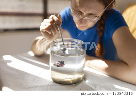 Woman feeding betta fish in glass jar with spoon 123843316