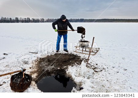 Using pole basket, man mines natural resources sapropel from lake floor, loading putrefaction into container, delivering organic fertilizer on sled for rural use. Using pole basket, man mines natural resources sapropel from lake floor, loading putrefaction into container, delivering organic fertilizer on sled for rural use. 123843341