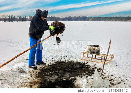Farmer extracts lake silt sapropel using basket on pole via ice hole for agriculture, transferring organic material to sled, enriching soil naturally. 123843342