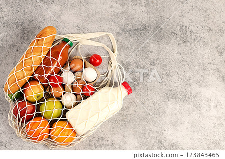 Fabric string bag with various natural products. Milk, eggs, baguettes, natural juice and fruits in a cotton bag on a concrete background, food delivery concept, advertising for a store. Fabric string bag with various natural products. Milk, eggs, baguettes, natural juice and fruits in a cotton bag on a concrete background, food delivery concept, advertising for a store. 123843465