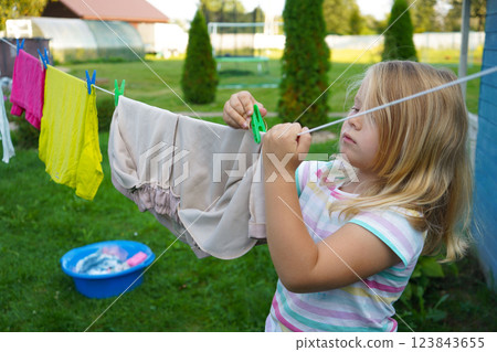 Wearing a cheerful summer dress, a six-year-old girl stretches her small arms to hang a damp shirt, her face full of focus. 123843655
