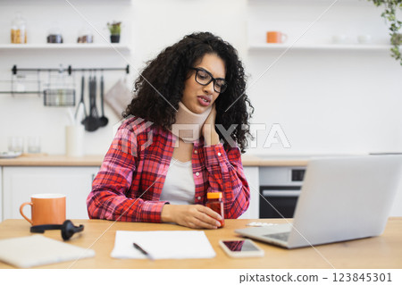 Woman of African descent suffers neck pain, consults healthcare professional online via laptop in kitchen environment, illustrating modern telemedicine solutions. 123845301