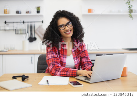 Smiling African woman with curly hair, wearing red shirt, working on laptop, at home kitchen. Scene conveys comfort, productivity, and warmth. 123845312