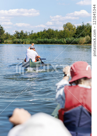 Man with two children enjoy having fun paddling canoe rent across lake river water wearing bright orange life vests hot sunny summer day. Healthy recreational activity lifestyle vacation trip journey 123845344