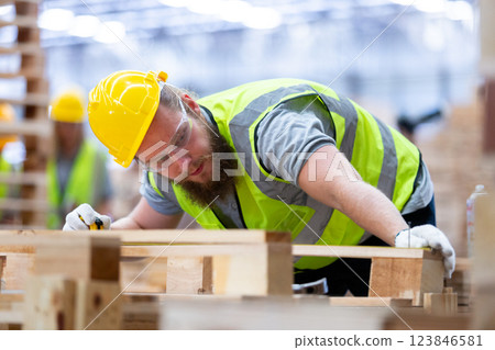 Wood factory construction worker wearing a yellow hard hat and safety vest focuses on measuring and crafting wooden materials in well-lit workshop. 123846581