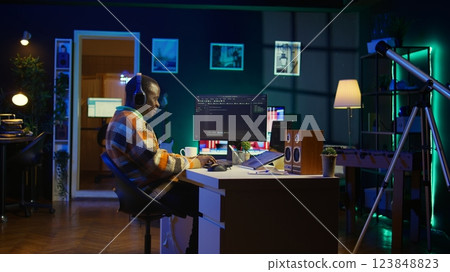 Black programmer remotely working in home office writing code on computer at his desk. Programming engineer IT expert in apartment typing lines of code at workstation, teleworking. Camera B. 123848823
