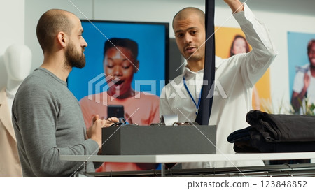 Retail associate presenting all models of ties for a young man shopper, client asking for formal accessories to match his suit. Store employee offers assistance for inspiration. Camera A. 123848852