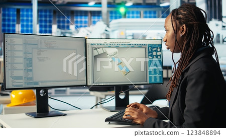 Programmer in photovoltaics factory using PC to update software of robot arms placing solar panels on assembly lines. Woman coding on computer, upgrading solar plant machinery, camera A 123848894