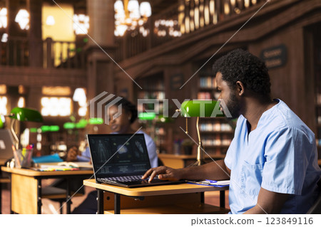 Black med school scholar studying and taking notes using online information, reviews a thesis project in the campus library. Young adult collecting academic data for medical research. Black med school scholar studying and taking notes using online information, reviews a thesis project in the campus library. Young adult collecting academic data for medical research. 123849116
