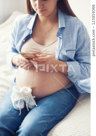A pregnant woman is seen resting comfortably on bed, holding a pair of baby shoes near her belly. She appears content and serene, enjoying a quiet moment 123859366
