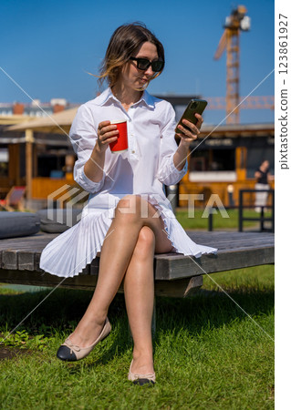 Woman in white dress wearing sunglasses and ballet flats, looking at smartphone while holding cup 123861927