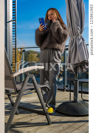 Woman on a sunny terrace in autumn Prague, holding coffee and smartphone in a grey faux fur coat Woman on a sunny terrace in autumn Prague, holding coffee and smartphone in a grey faux fur coat 123861983