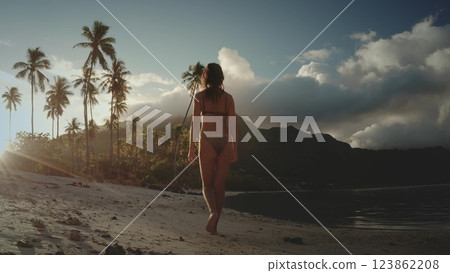 A woman walks barefoot on a white sand beach in French Polynesia, with a line of palm trees and a mountain in the background. The sun is setting, casting a golden glow on the scene. 123862208