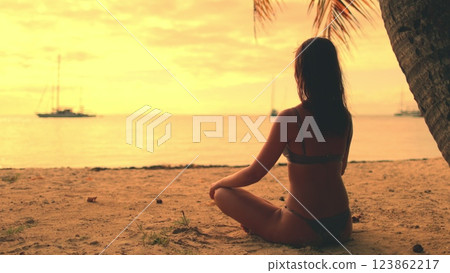 Young woman is enjoying a moment of peace and tranquility as she practices yoga on a secluded beach at sunset. The warm glow of the setting sun creates a serene and magical atmosphere 123862217