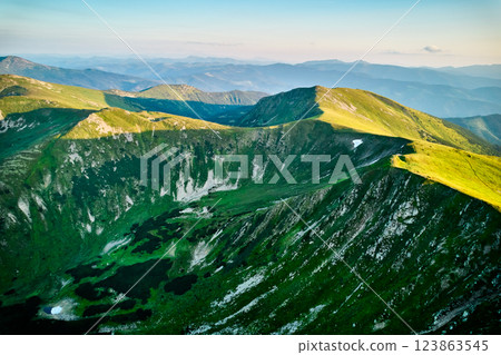 Aerial view of rugged mountain range with lush green slopes at sunset. Landscape showcases beauty of untouched nature with distant peaks and valleys bathed in soft sunlight. Carpathians, Chornogora. 123863545