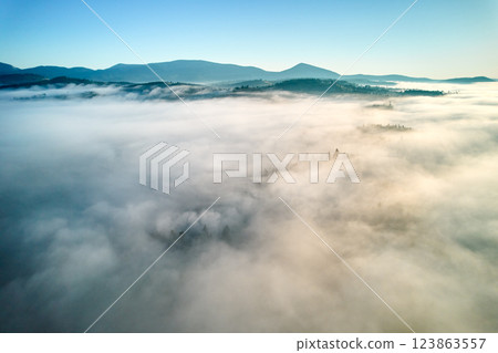 Aerial view of landscape enveloped in thick, rolling clouds. Verdant hills and scattered trees barely peek through, while backdrop of distant mountains completes this serene and dreamlike scene. Aerial view of landscape enveloped in thick, rolling clouds. Verdant hills and scattered trees barely peek through, while backdrop of distant mountains completes this serene and dreamlike scene. 123863557