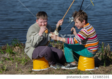 The boys are friends on a fishing trip by the river eating a bun and laughing. Children are friends on a summer fishing trip. 123865101
