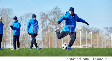 Strength in motion. Football player in blue jacket striking ball with controlled power, teammates watching training progression. Strength in motion. Football player in blue jacket striking ball with controlled power, teammates watching training progression. 123865948