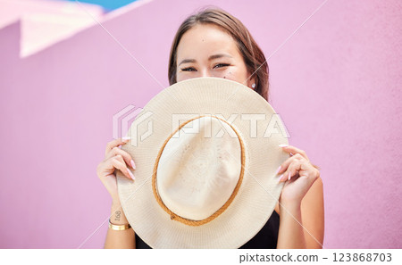 Travel, fashion and summer hat with a woman tourist in the city on a pastel color pink wall background. Happy, holiday and tourist with a female traveler covering her face with a sunhat in town 123868703