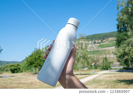 Man holding in the hand white thermo stainless bottle with refreshing water on the nature. 123868907